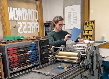 A woman holds a sheet of paper over a printing press in a studio filled with printing materials. On the window behind her there is text that reads, "COMMON PRESS."