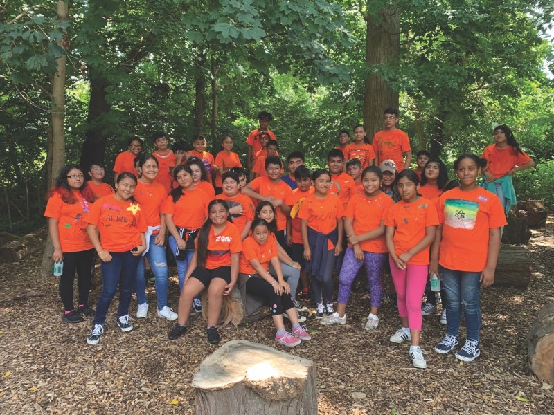 A photo of a large group of Latinx children in the woods all wearing orange ACLAMO t-shirts