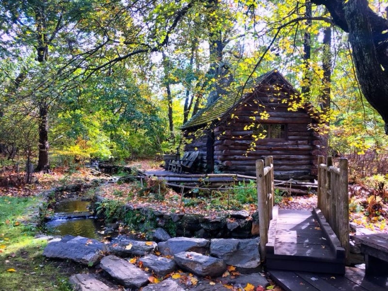 A log cabin sits along a creek with a wooden bridge.