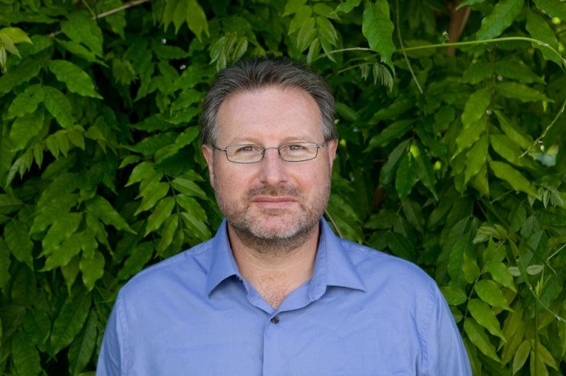 A man in a blue shirt stands in front of bright green foliage.
