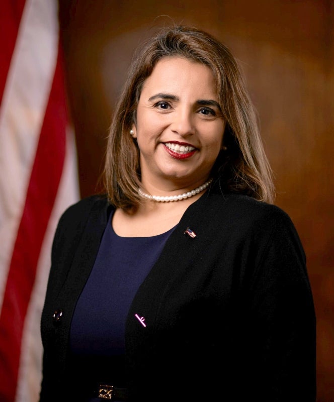 A Latina woman smiles at the camera with the American flag off to the side.