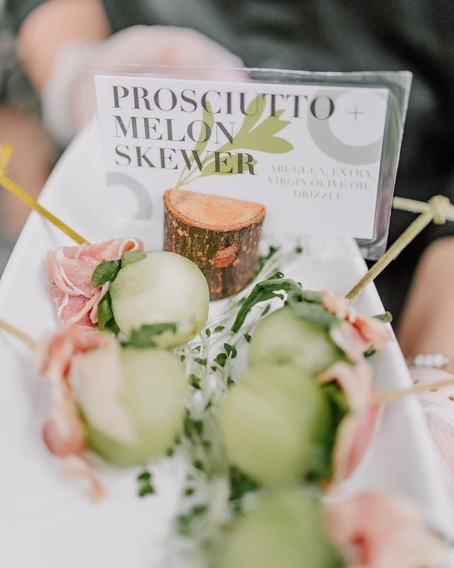 A plate of appetizers with a sign that reads "Prosciutto Melon Skewer."