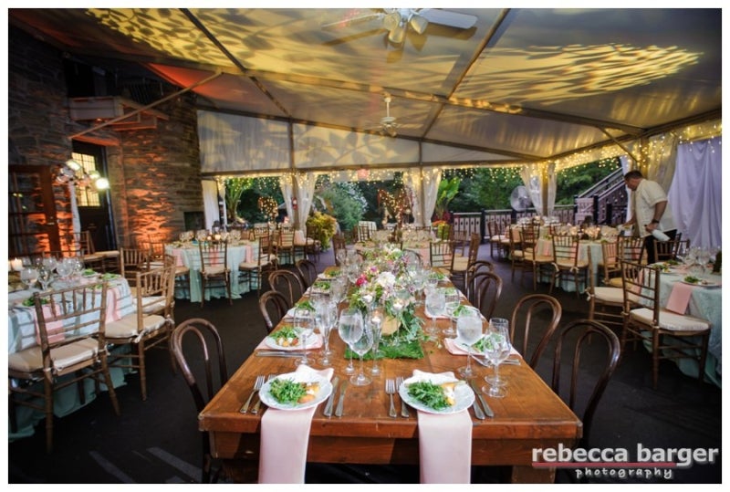 A reception setup of tables, chairs, place settings, and flowers under a white tent. 