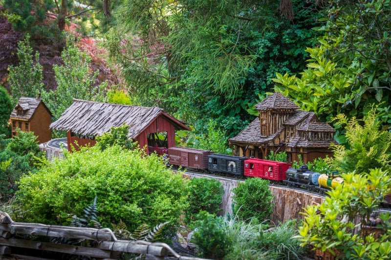 A model train rides through a miniature covered bridge surrounded by greenery. 