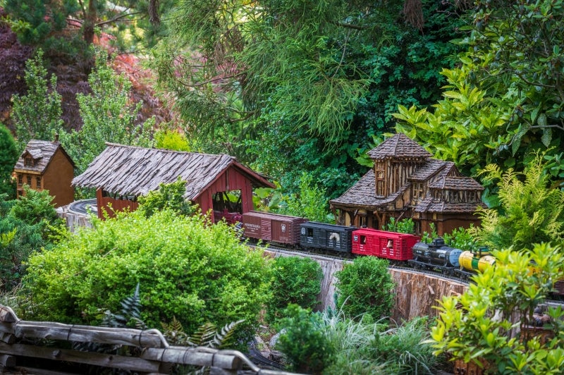 A model train rides through a miniature covered bridge surrounded by greenery.
