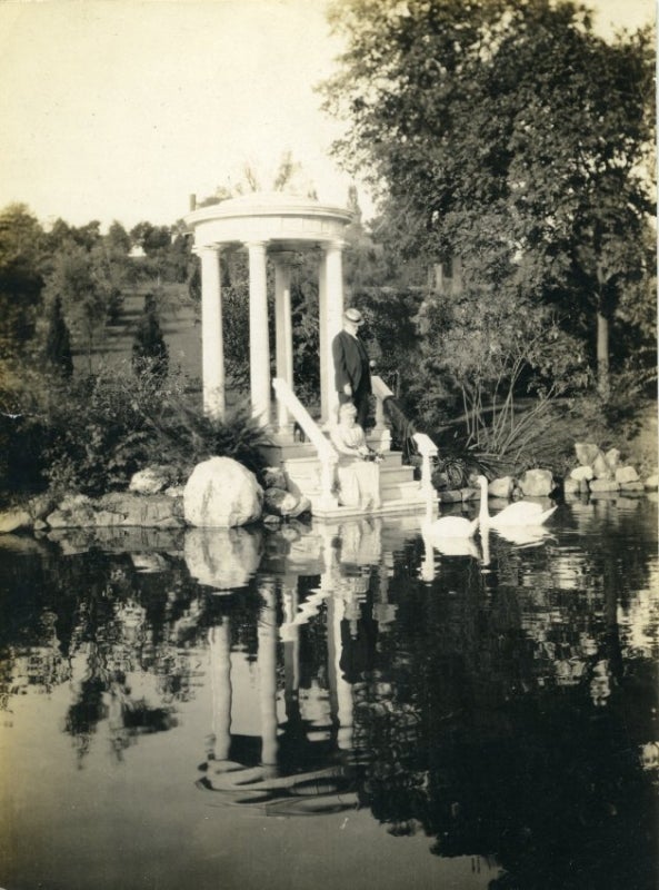 A black and white photograph of a man and women near the edge of a pond looking at two swans.