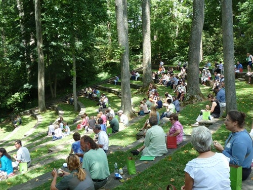 A group of people sit on a sloped hill in the trees. 