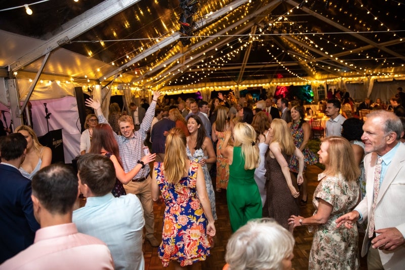 A large group of people in informal attire dance under a clear-top tent with string lights.