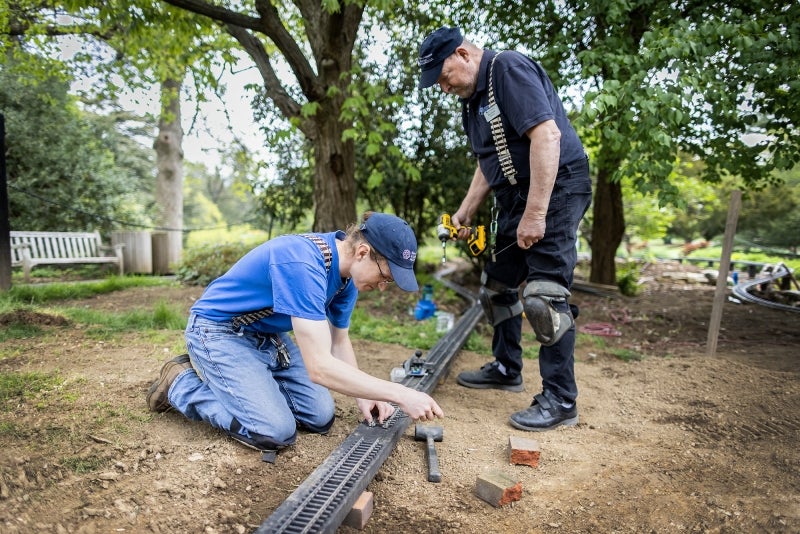 Two men work on a miniature railroad track in a public garden. 