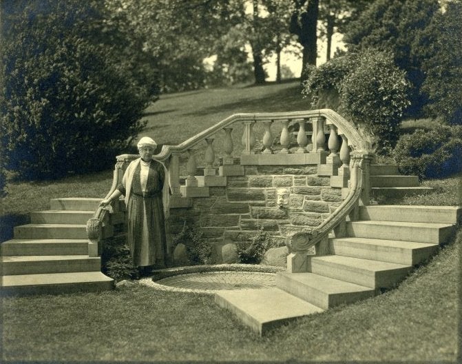 A sepia-toned photograph from the 1920s of a woman white white hair standing outside in front of stairs.