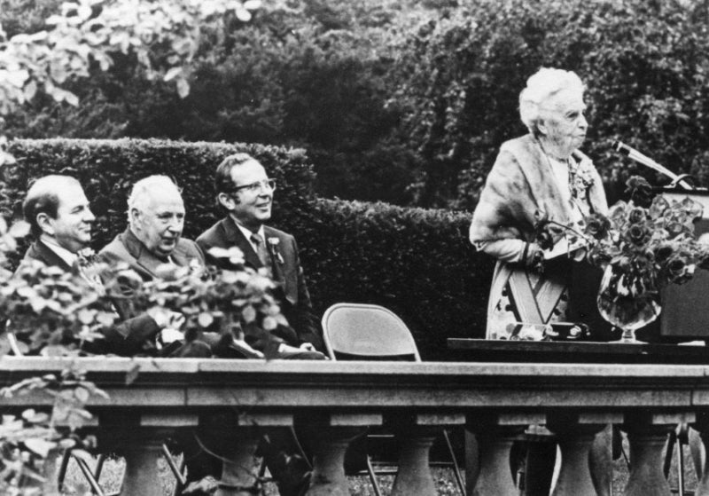 A black-and-white photos of a woman with white hair speaking at a podium with three men smiling behind her.