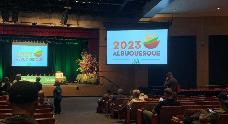 A dimly lit auditorium with people both sitting and standing, and a projector that reads, "2023 Albuquerque ISA."