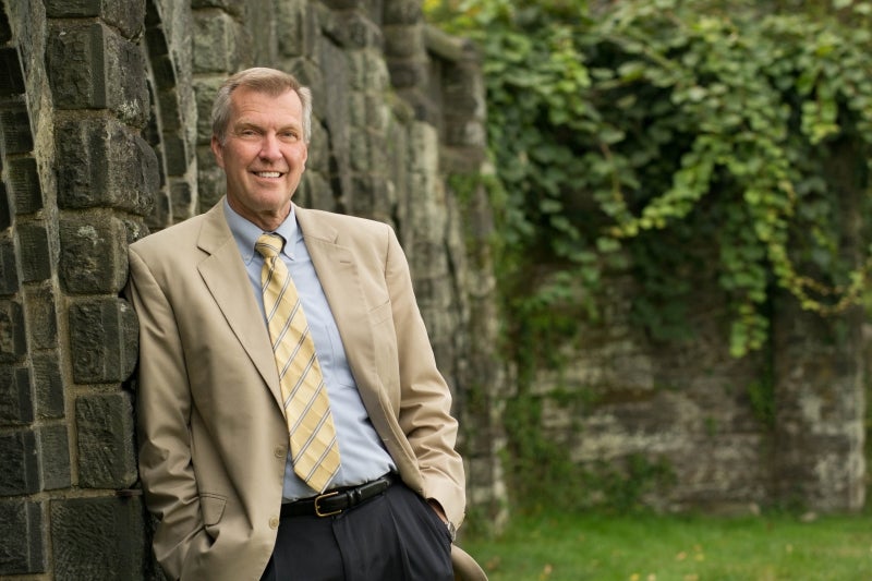 A man leans against a stone wall posing for the camera with greenery in the background.