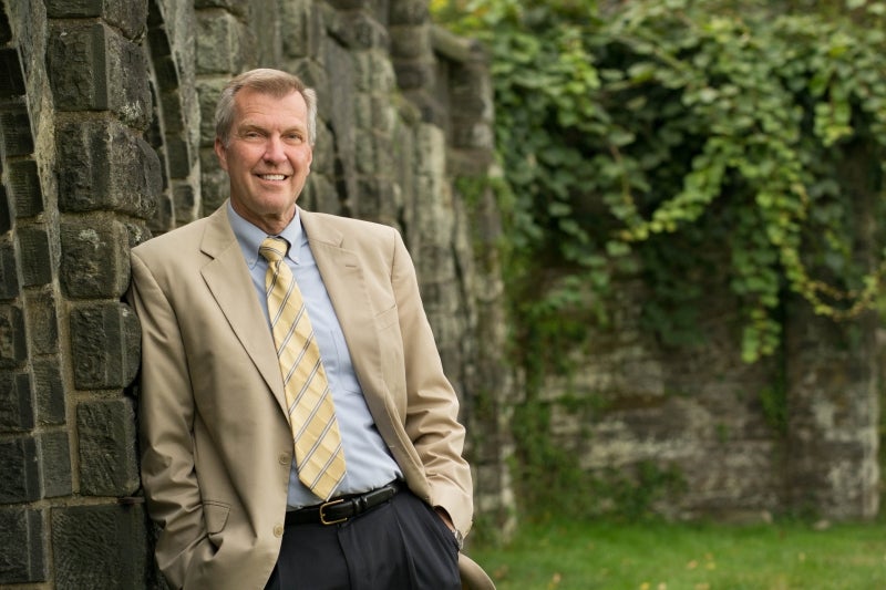 A man leans against a stone wall posing for the camera with greenery in the background. 