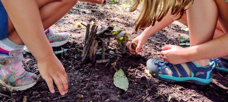 Two young children build a small house out of sticks and leaves.