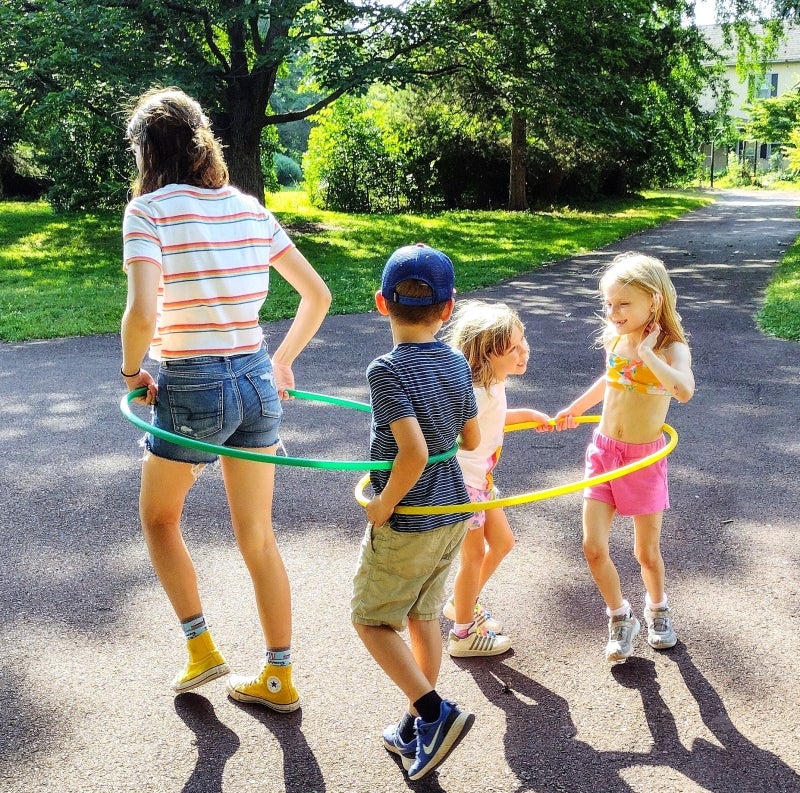 A young adult shows three small children how to hula hoop outdoors.
