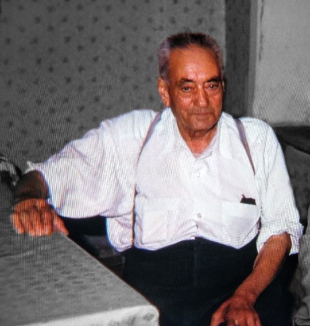 An older man in a white shirt and suspenders sits in a chair.