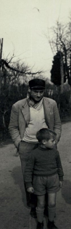 A man in a hat and jacket stands with a young boy in a sweater and shorts.