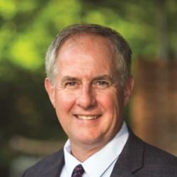 A headshot of a man with gray hair smiling outdoors in a suit.