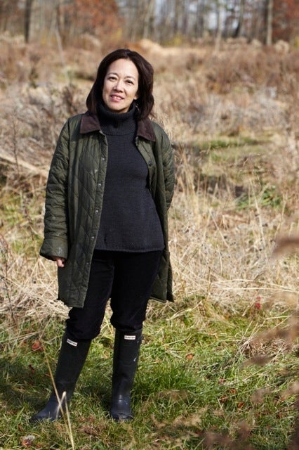 Woman with black hair wearing a dark coat and black boots standing in a field of brown weeds