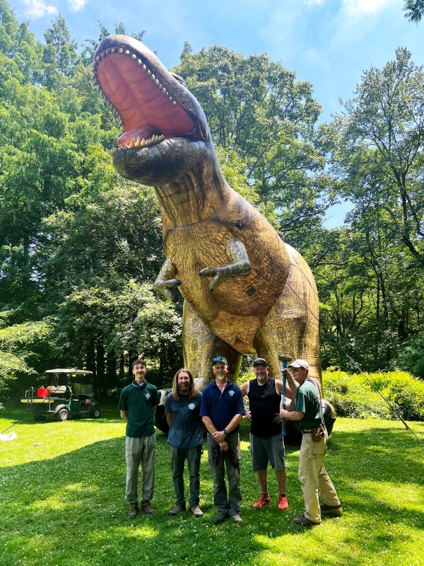 Five men pose in front of a 25-foot inflatable Tyrannosaurus rex outdoors in a green space. 
