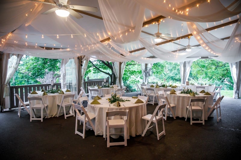 The inside of an outdoor events tent with round tables and chairs with table settings and décor, and string lights and fabric on the ceiling.