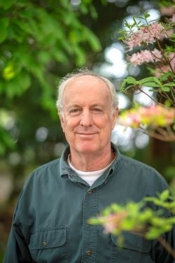 A headshot of a man wearing a dark green button up shirt stand next to a bloom plant.