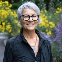 Headshot of a woman with short grey hair, black-framed glassed, and a black shirt standing outdoors in a blooming garden.
