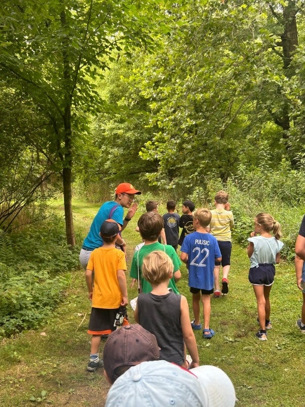 The backs of a group of children walking through a green wooded area with an adult talking to them. 