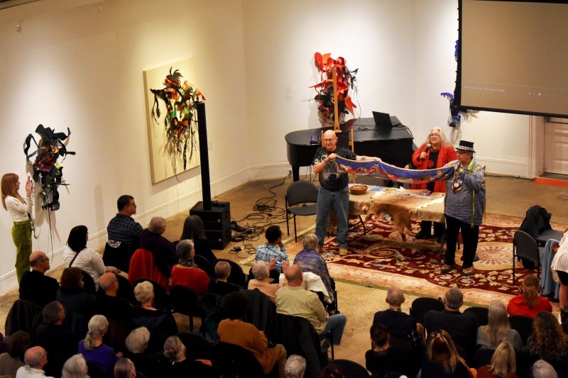 A seated audience watches two members of the Lenape Nation of PA hold up a long piece of beaded fabric while a third speaks on a microphone.