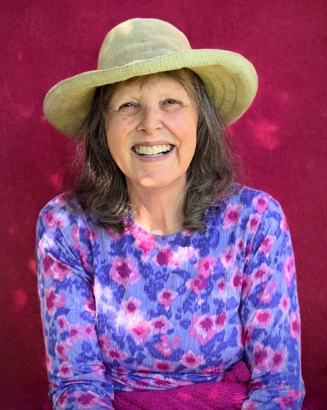 Smiling white woman in a purple and pink long-sleeve shirt and wearing a beige brimmed hat