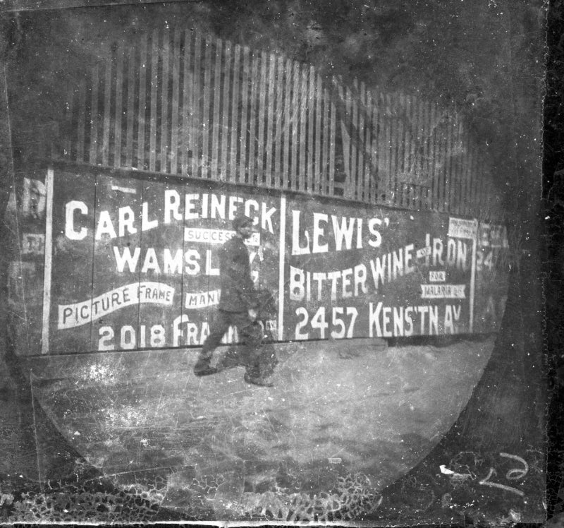 A black-and-white photographs from the late 1800s of a man walking along a building will business signs.