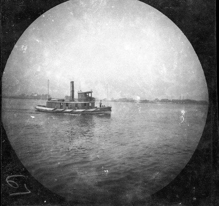 A black-and-white photograph of a tugboat on a body of water. 