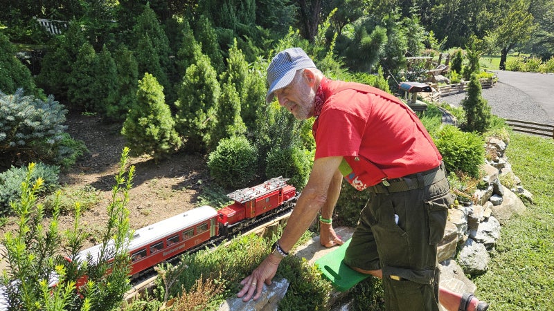 Man in red t-shirt and blue and white striped train engineer hat leans over a red train car on a track