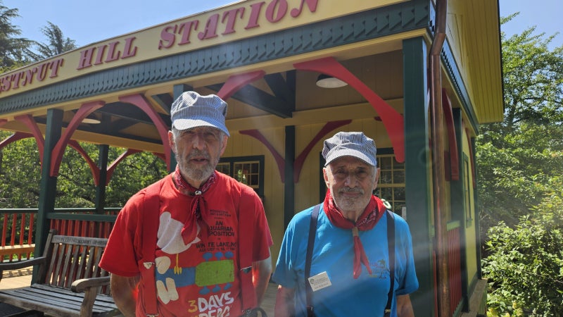 Two older man in conductor caps and suspenders stand in front of a fabricated train station in a model train display in a public garden. 