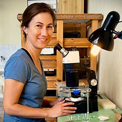 A headshot of a women with dark hair and a blue t-shirt in a laboratory smiling at the camera and sitting in front of a microscope.  