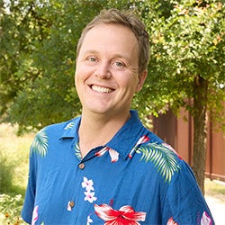 A headshot of a smiling man outdoors with blonde hair and a blue Hawaiian shirt. 