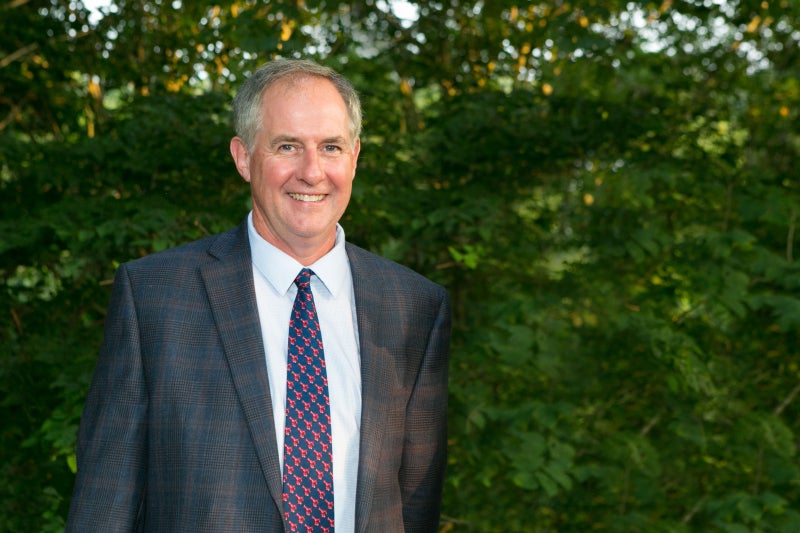 White man with gray hair wearing a dark gray suit, white shirt, and purple and blue tie