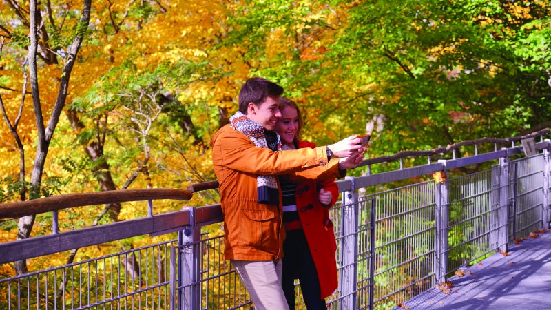 Two people lean against a fence in front of a scene of fall foliage and take a selfie. 