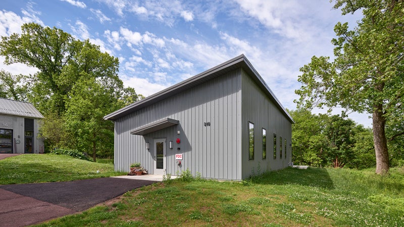 A small grey building with a slanted roof and windows stands in an outdoor green space. 